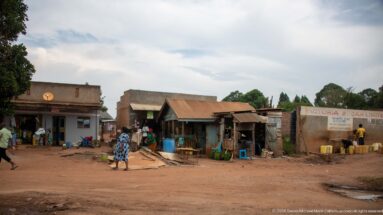 Row of simple timber, metal, and masonry shops along a dirt road with people walking between open-front businesses.