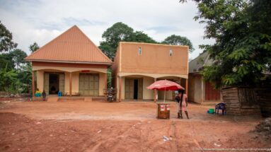 Small Ugandan roadside shops and an umbrella-covered kiosk facing a broad unpaved red-earth street.