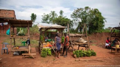 Two people beside a simple roadside stall with green banana bunches and assorted produce on a dirt forecourt.