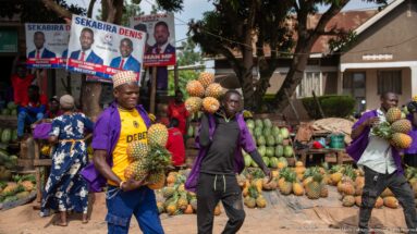 Men carrying multiple pineapples past a roadside market piled with pineapples, watermelons, and other produce.
