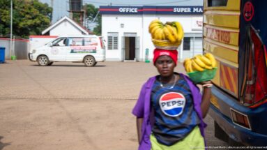 Woman balancing yellow bananas on her head and carrying more fruit beside a large vehicle at a roadside stop.