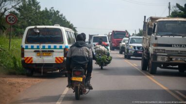 Motorcycles riding along the edge of a busy two-lane highway beside vans and trucks, with a banana-laden motorcycle ahead.