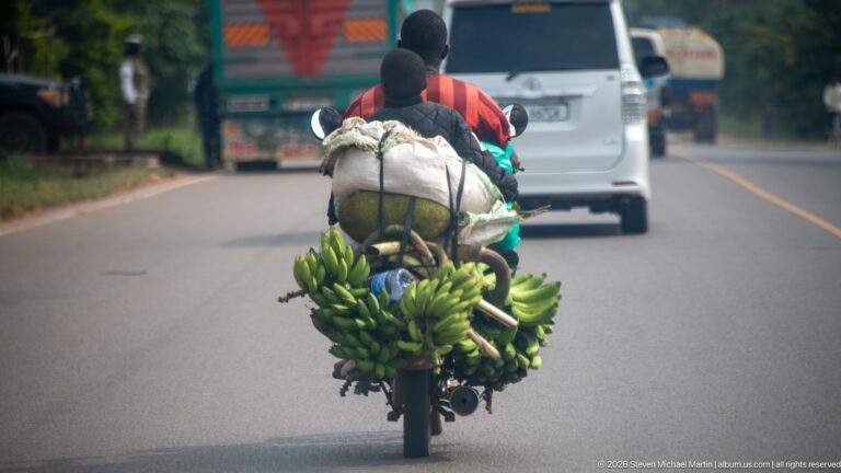 Motorcycle carrying large bunches of green bananas and sacks along a paved Ugandan highway behind other vehicles.