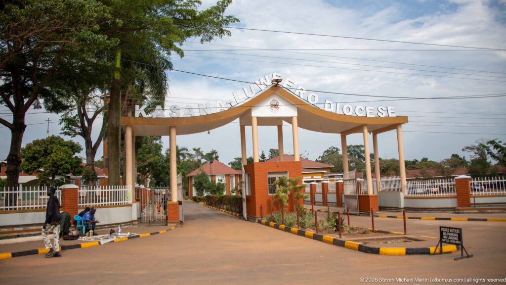 Arched entrance gate reading Kasana-Luweero Diocese, opening onto a landscaped drive and red-roofed buildings.