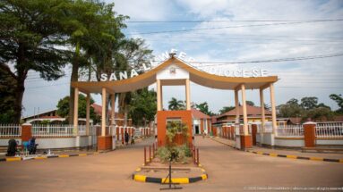 Centered view of the Kasana-Luweero Diocese entrance arch, driveway, landscaped median, fencing, and red-roofed buildings.