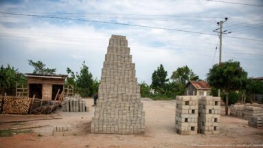 Tall stacks of concrete masonry blocks in an open roadside yard with timber piles and a simple work shelter.