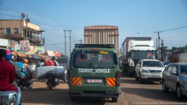 Green tourist vehicle surrounded by motorcycles, cars, and trucks in dense traffic on a busy Ugandan road.