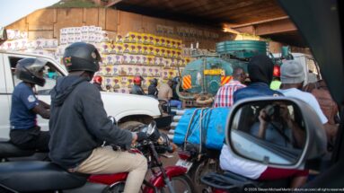 Motorcyclists in helmets beside a roadside stall packed with colorful goods, seen from inside a vehicle in traffic.