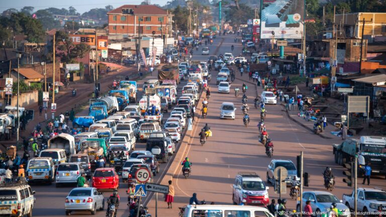 Heavy traffic of minibuses, cars, motorcycles, and pedestrians filling a broad commercial road in a Ugandan city.