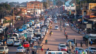 Heavy traffic of minibuses, cars, motorcycles, and pedestrians filling a broad commercial road in a Ugandan city.