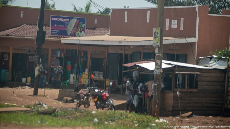 Motorcycles parked outside small roadside shops with pedestrians gathered under a corrugated awning on a red-earth verge.