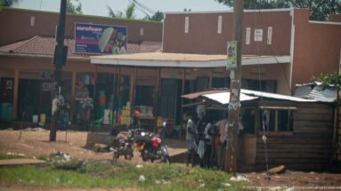 Motorcycles parked outside small roadside shops with pedestrians gathered under a corrugated awning on a red-earth verge.