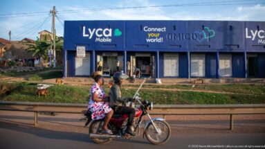 Motorcycle carrying two people past blue mobile-phone shops, with goats grazing on the roadside verge.