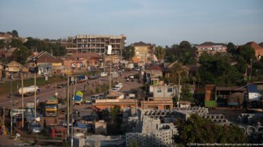 Busy Ugandan commercial road with minibuses, trucks, motorcycles, shops, utility lines, and stacks of concrete construction pieces.