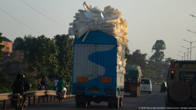Blue box truck driving in traffic with a towering irregular load of white sacks stacked above its cargo body.