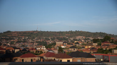 Hillside neighborhood with dense red-roofed houses rising toward a ridge with communication towers under a clear blue sky.