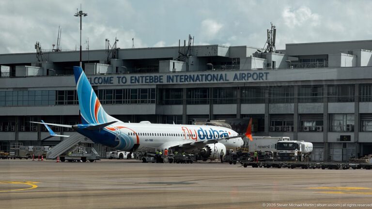 Flydubai jet parked beside the terminal at Entebbe International Airport with ground vehicles and service equipment on the apron.