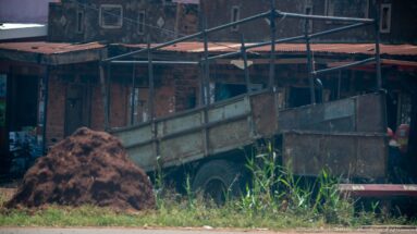 Weathered open metal trailer beside a mound of reddish-brown organic material and brick roadside workshops.