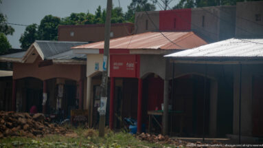 Low painted roadside shops with shaded fronts, utility poles, and piles of brown goods or produce outside one bay.