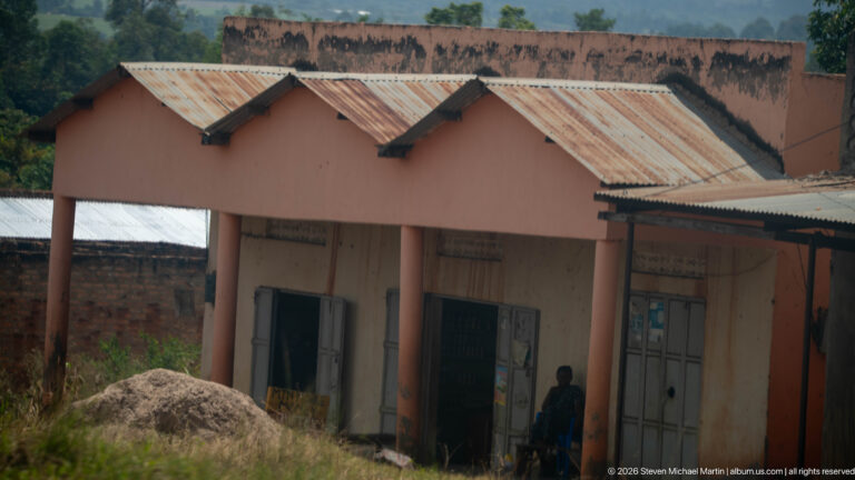 Pink masonry shop building with repeated peaked roof bays, corrugated roofing, and metal doors along a roadside.