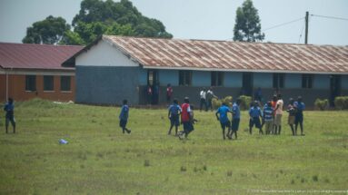 Schoolchildren playing and gathering on a grassy field in front of long low classroom buildings.