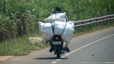 Motorcyclist riding beside a guardrail with two huge white sacks strapped across the rear of the bike.