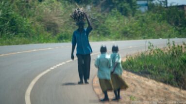 Man carrying a bulky bundle on his head while two schoolchildren walk along the shoulder of a curving paved road.