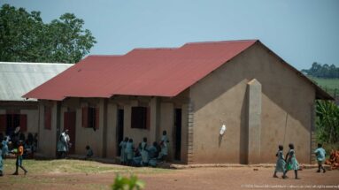 Children in school uniforms gathered outside a simple masonry classroom building with a red metal roof.