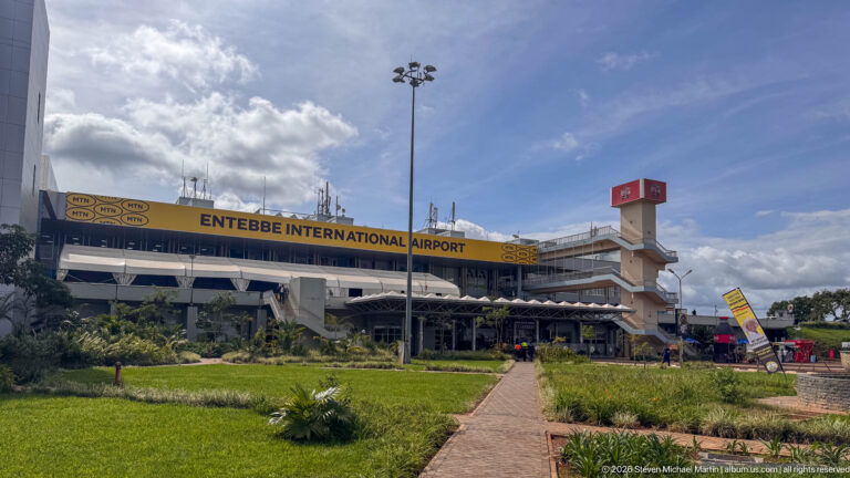 Entebbe International Airport terminal behind landscaped lawns and a paved walkway under a bright blue sky.