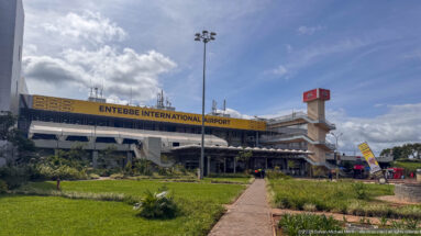 Entebbe International Airport terminal behind landscaped lawns and a paved walkway under a bright blue sky.