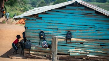 Child beside a blue-painted timber building with several backpacks hanging from the wall under a corrugated roof.