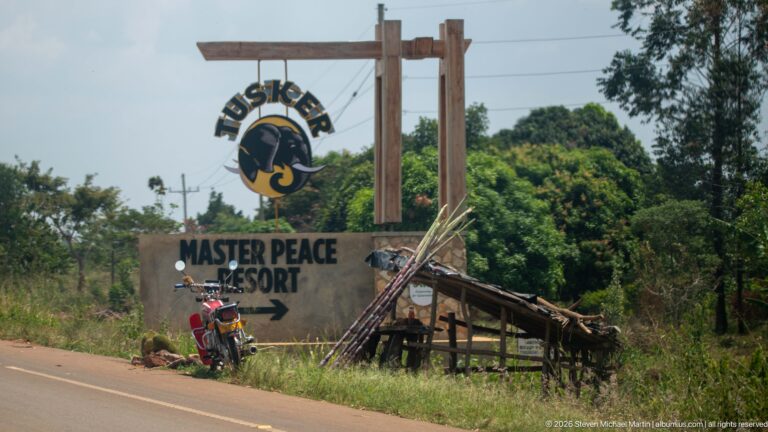 Tusker-branded Master Peace Resort sign with directional arrow beside a motorcycle and rough timber shelter.