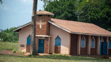 Small brick-and-plaster church with a white cross, pointed-arch windows, rust-colored roof, and grassy yard.