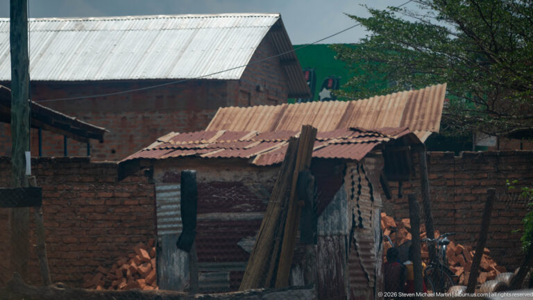 Small weathered corrugated-metal shed with a rusted roof, stacked poles, and brick houses behind.