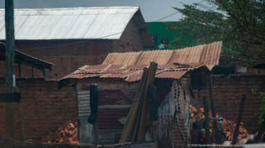 Small weathered corrugated-metal shed with a rusted roof, stacked poles, and brick houses behind.