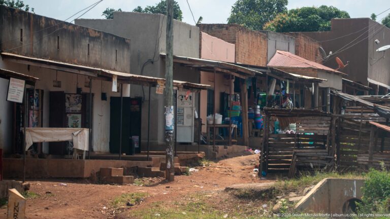 Small open-front shops and timber fencing line a narrow red-earth lane with merchandise and posters outside.