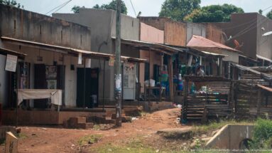 Small open-front shops and timber fencing line a narrow red-earth lane with merchandise and posters outside.
