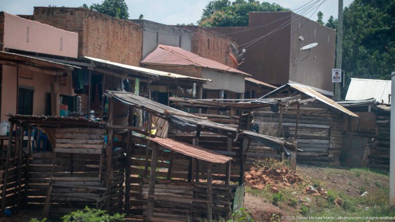 Timber roadside stalls with corrugated roofs stand before brick buildings, utility wires, satellite dishes, and red-earth ground.
