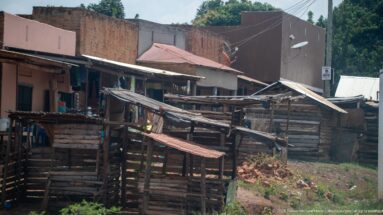 Timber roadside stalls with corrugated roofs stand before brick buildings, utility wires, satellite dishes, and red-earth ground.