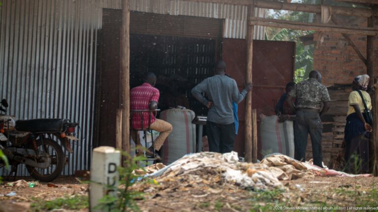 People standing around large white sacks inside an open corrugated-metal and brick warehouse with a motorcycle beside the entrance.