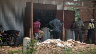 People standing around large white sacks inside an open corrugated-metal and brick warehouse with a motorcycle beside the entrance.