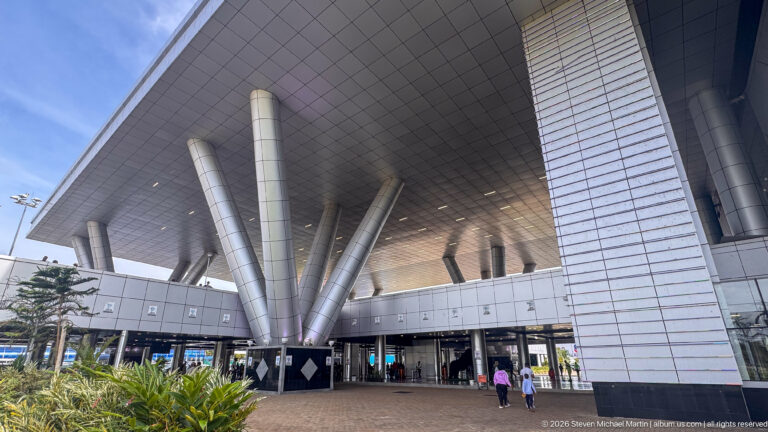 Large airport canopy supported by angled silver columns above glass entrances and landscaped planting at Entebbe International Airport.