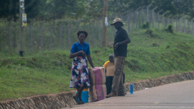 Woman standing beside tall rolled or bundled woven goods with a man and child at the edge of a paved road.