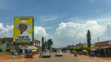 Large yellow Yoweri Museveni campaign billboard above a busy commercial street with cars, motorcycles, stalls, and umbrellas.