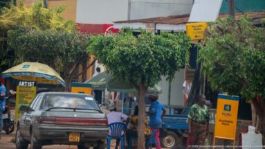 Yellow MoMo 'Available Here' sign beside a parked car, plastic chairs, umbrellas, pedestrians, and shaded roadside shops.