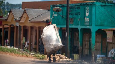 Man walking beside a paved commercial street with an oversized white sack carried over his shoulder and back.