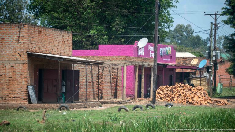 Unfinished brick shops beside a pink Family Soap building, brick pile, satellite dishes, grass, and utility poles.