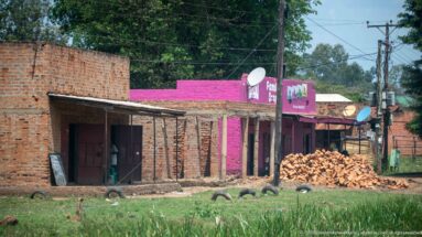 Unfinished brick shops beside a pink Family Soap building, brick pile, satellite dishes, grass, and utility poles.
