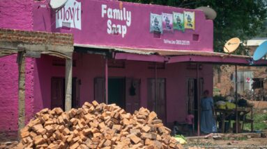 Pink Family Soap storefront behind a large pile of red bricks, with satellite dishes and people near an adjoining stall.