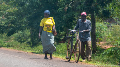 Woman walking beside a man pushing a bicycle along the grassy shoulder of a paved road.